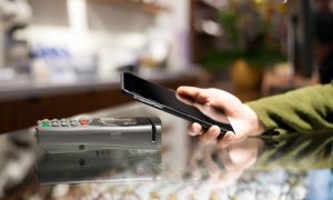 Imagen en la que se muestra el mostrador de un local, y sobre este una TPV y el brazo de una persona que aproxima su móvil para pagar con él.