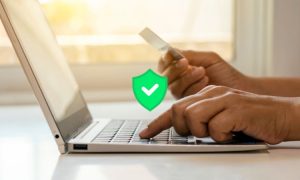 Imagen de un pc con una manos sobre su teclado, la persona está haciendo un pago online ya que se ve cómo coge con su mano derecha una tarjeta de débito.