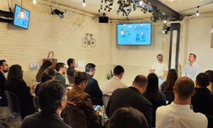 Personas sentadas atienden a dos presentadores de pie en una sala, con pantallas mostrando presentaciones en la pared. Ambiente de conferencia.