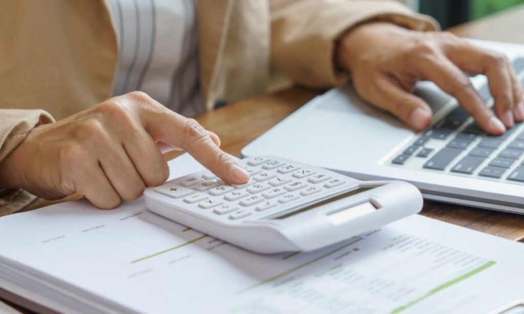 La imagen muestra a una persona realizando cálculos financieros o contables. Está utilizando una calculadora y un ordenador portátil, mientras revisa documentos con gráficos y cifras sobre una mesa. La escena transmite una sensación de análisis y gestión económica, posiblemente relacionada con la administración de costes o la planificación presupuestaria.