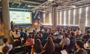 La imagen muestra una sala llena de asistentes siguiendo una ponencia en un evento profesional. Una persona está en el escenario explicando un diagrama proyectado en la pantalla, mientras el público, compuesto por unas cuarenta personas, escucha con atención. El ambiente es moderno e informal, con luz natural entrando por los ventanales y decoración urbana en las paredes, transmitiendo un clima de aprendizaje y networking propio de un encuentro de marketing o SEO.