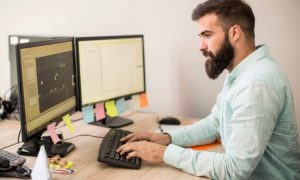 La imagen muestra a un hombre joven con barba sentado frente a una computadora de escritorio con dos pantallas. Está concentrado, escribiendo al teclado mientras mira fijamente a la pantalla.