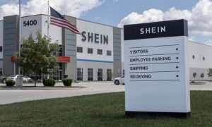 Edificio de oficinas con letrero de entrada en primer plano que dice "SHEIN", señalando áreas de VISITANTES, APARCAMIENTO EMPLEADOS, ENVÍOS y RECEPCIÓN. Bandera americana ondeando.