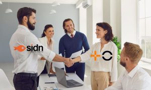 un grupo de cinco personas en un entorno profesional, probablemente en una sala de reuniones. Dos de ellas —un hombre a la izquierda y una mujer a la derecha— se están dando la mano, simbolizando un acuerdo o colaboración. Ambos visten de manera formal, y sus compañeros, también vestidos de forma profesional, los observan sonrientes. Sobre la imagen aparecen los logotipos de SIDN (en el lado izquierdo) y de MasOrange (en el derecho), reforzando la idea visual de una alianza entre ambas compañías. La escena transmite profesionalismo, cooperación y cierre de acuerdos estratégicos.