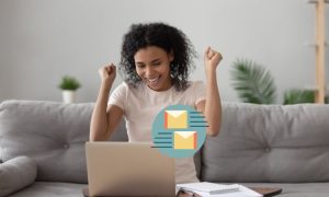 Persona celebrando recibir correos en un portátil, sentada en un sofá gris con plantas de fondo. Éxito en marketing digital desde casa.