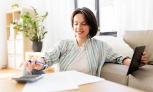 una mujer sentada cómodamente en un sofá, con un ambiente luminoso y relajado en su hogar. Sostiene un teléfono móvil en una mano mientras con la otra escribe en una hoja de papel con un bolígrafo. En la mesa frente a ella, se observan varias facturas y una calculadora, lo que sugiere que está trabajando en tareas financieras o revisando documentos importantes. Este tipo de escena refleja el tipo de enfoque que los negocios de ecommerce deberán adoptar para cumplir con la nueva normativa de facturación electrónica Verifactu, que entra en vigor el 1 de enero de 2026.