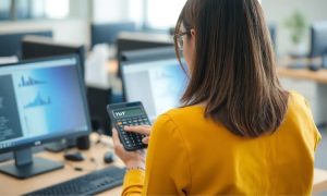 Persona revisa calculadora con texto "YoY" frente a dos pantallas de ordenador mostrando gráficos en una oficina moderna. Contexto empresario.