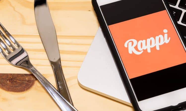 Teléfono mostrando el logo de Rappi, colocado sobre una mesa junto a cubiertos; ideal para marketing de aplicaciones de entrega y comida a domicilio.