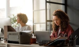 Dos personas trabajando en un entorno de oficina moderno. Una mujer está sentada en un escritorio, trabajando en una computadora portátil, tiene el cabello rizado y lleva una chaqueta roja. Al fondo, otra mujer está sentada de espaldas, trabajando en una computadora de escritorio. La oficina tiene luz natural que entra por grandes ventanas, y hay plantas y suministros de oficina sobre los escritorios. La atmósfera es enfocada y profesional.